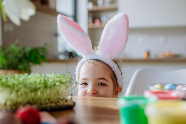 Wielkanocne godziny otwarcia w M1 Poznań. Dziewczynka w uszach zająca wielkanocnego, wychylająca się zza stołu.