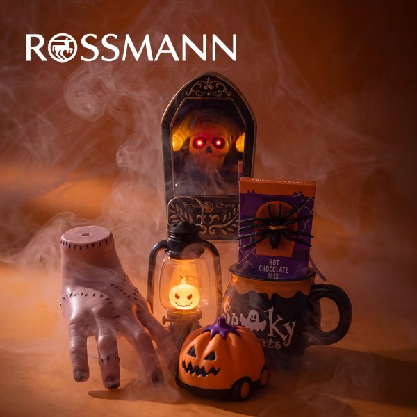 Dekoracje na Halloween na pomarańczowym tle spowitym mgłą: sztuczna ręka, dzwonek z czaszką o świecących czerwonych oczach, latarnia z podświetlaną dynią, pojazd w kształcie dyni oraz kubek z opakowaniem czekolady do picia. U góry białe logo Rossmann.
